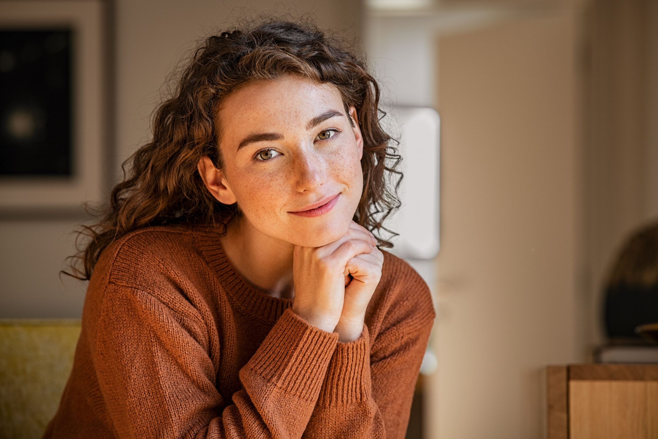 Portrait of satisfied young woman relaxing at home. Successful woman with hand on chin smiling and looking at camera. Beautiful natural girl stay at home with a serene expression.